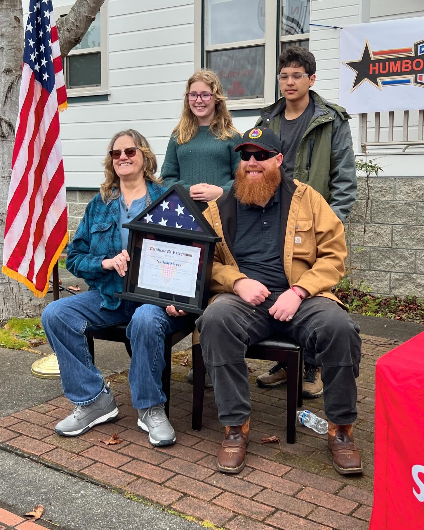 A Humboldt Heroes honoree seated with family and holding a framed flag and Certificate of Recognition